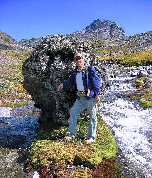 A man standing in front of a giant boulder in Greenland next to a stream of water.